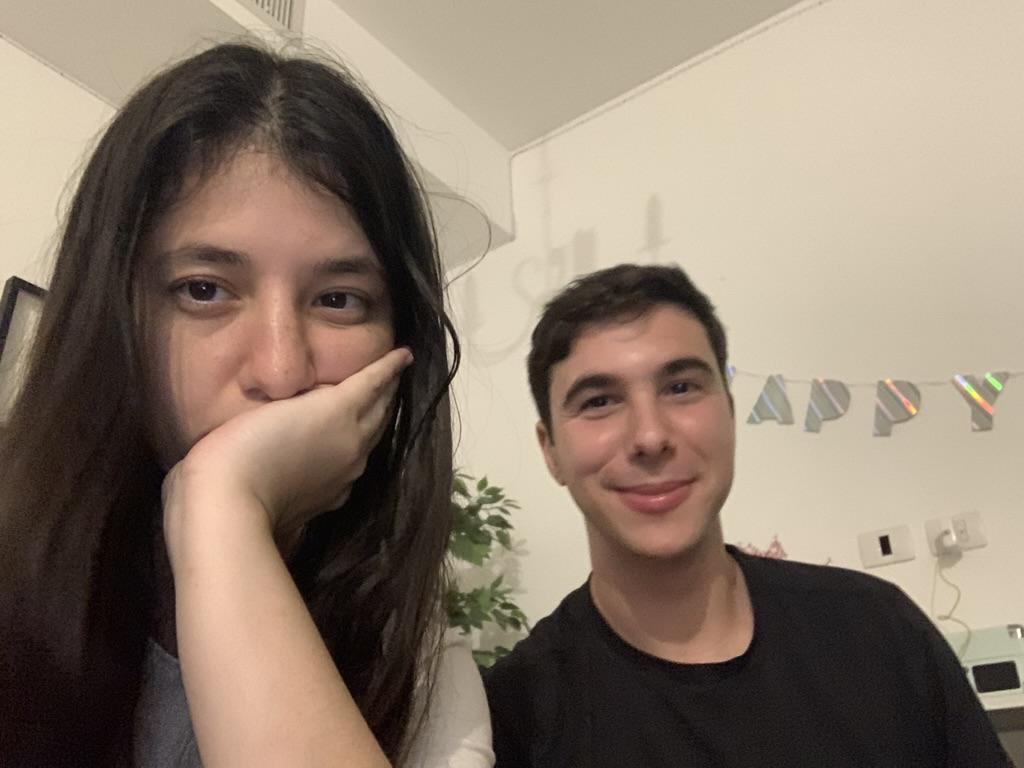 Hershel and Elia standing together in front of a HAPPY letter garland. Elia with hand near her face, Hershel smiling at the camera.