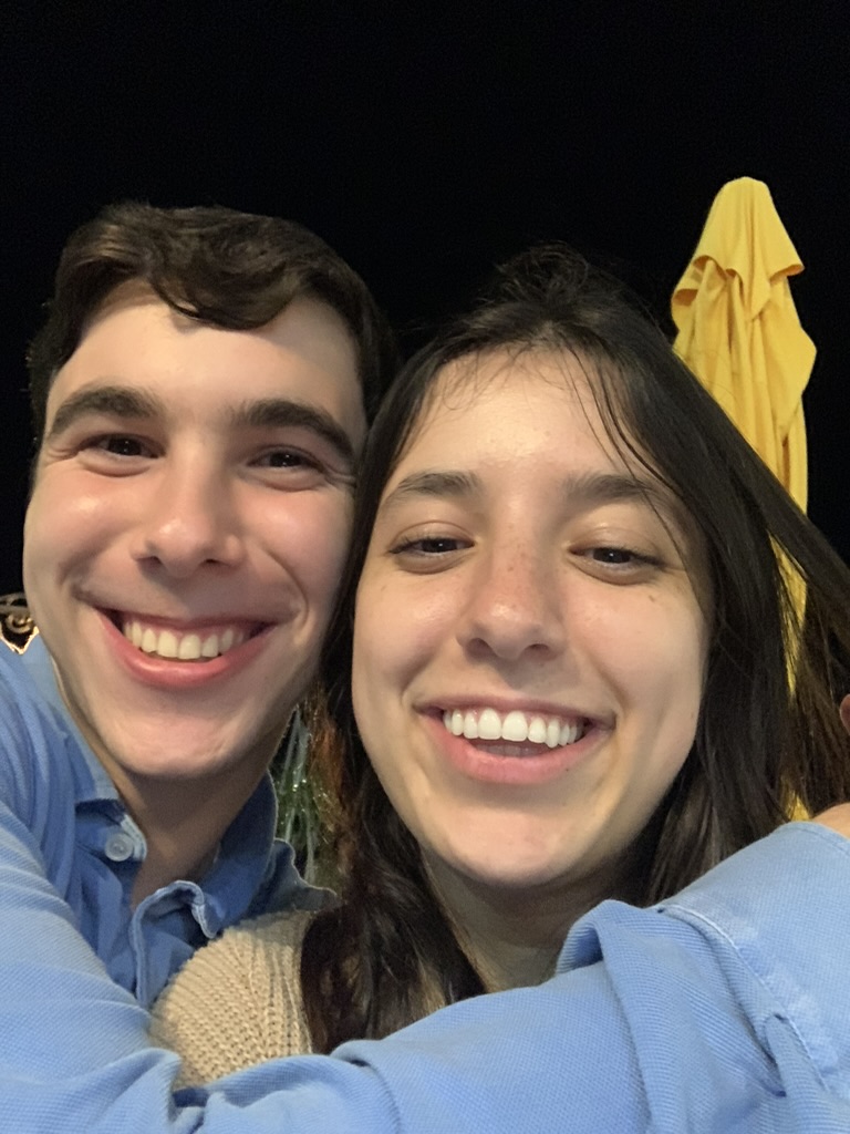 Hershel in blue button-down and Elia in light tan, posed and smiling together against a dark background.