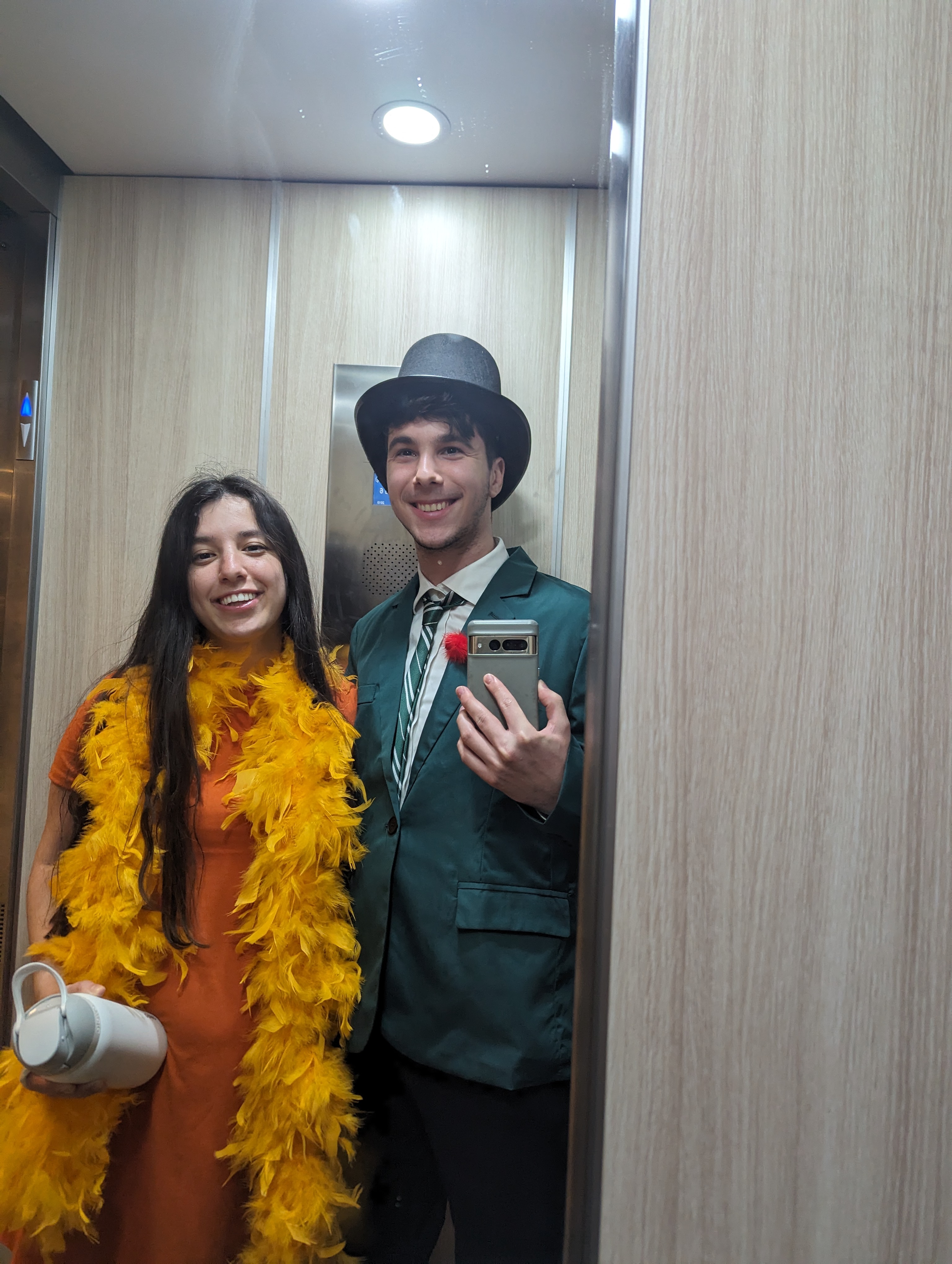 Elevator mirror selfie. Elia in orange dress with yellow feather boa. Hershel in green Slytherin suit jacket, striped tie, and black top hat.