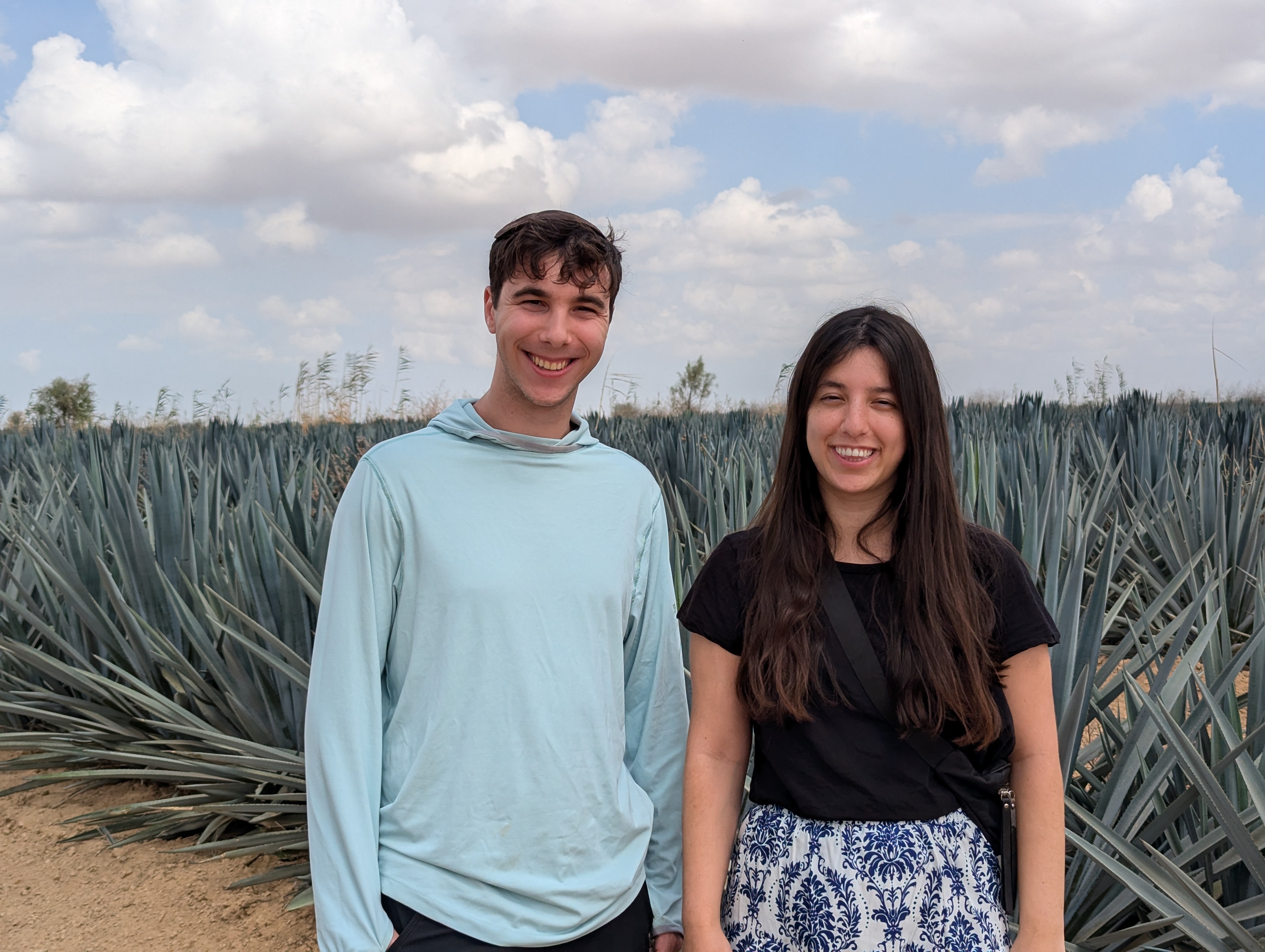 Hershel in light blue hoodie and Elia in black t-shirt with patterned blue-and-white skirt, standing in front of a field of blue agave plants under partly cloudy sky.
