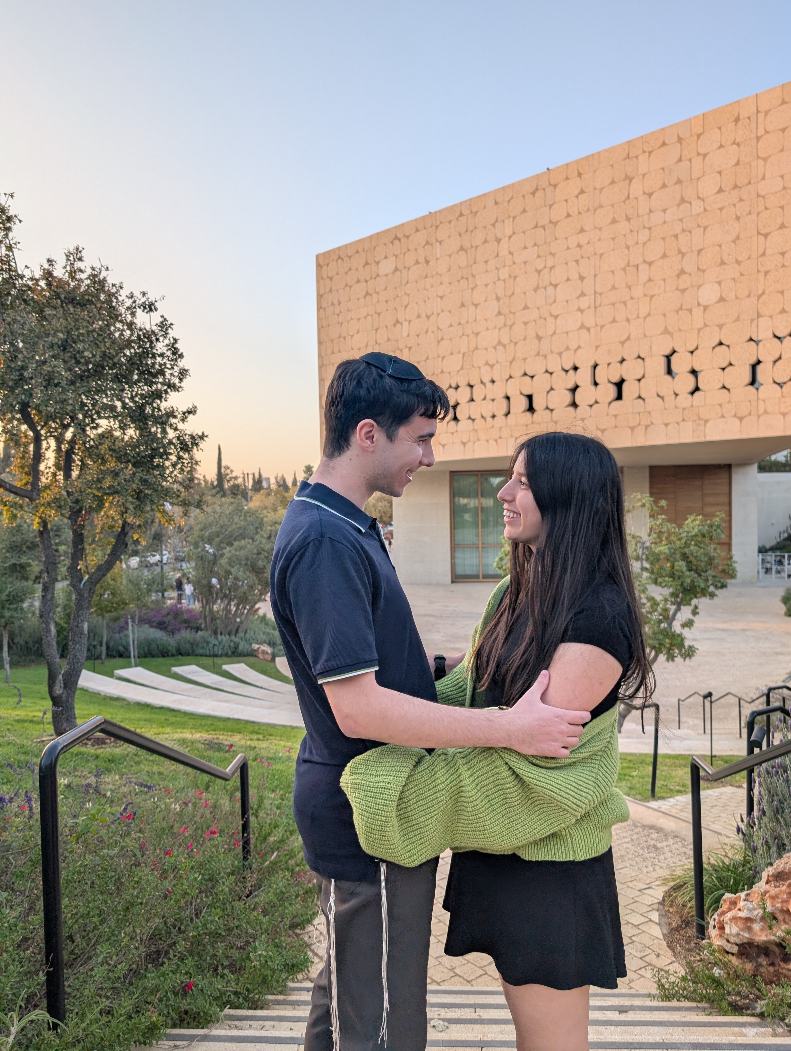 Hershel and Elia embracing on the front steps of the National Library of Israel in Jerusalem at golden hour, looking at each other with the building's geometric stone facade as backdrop.
