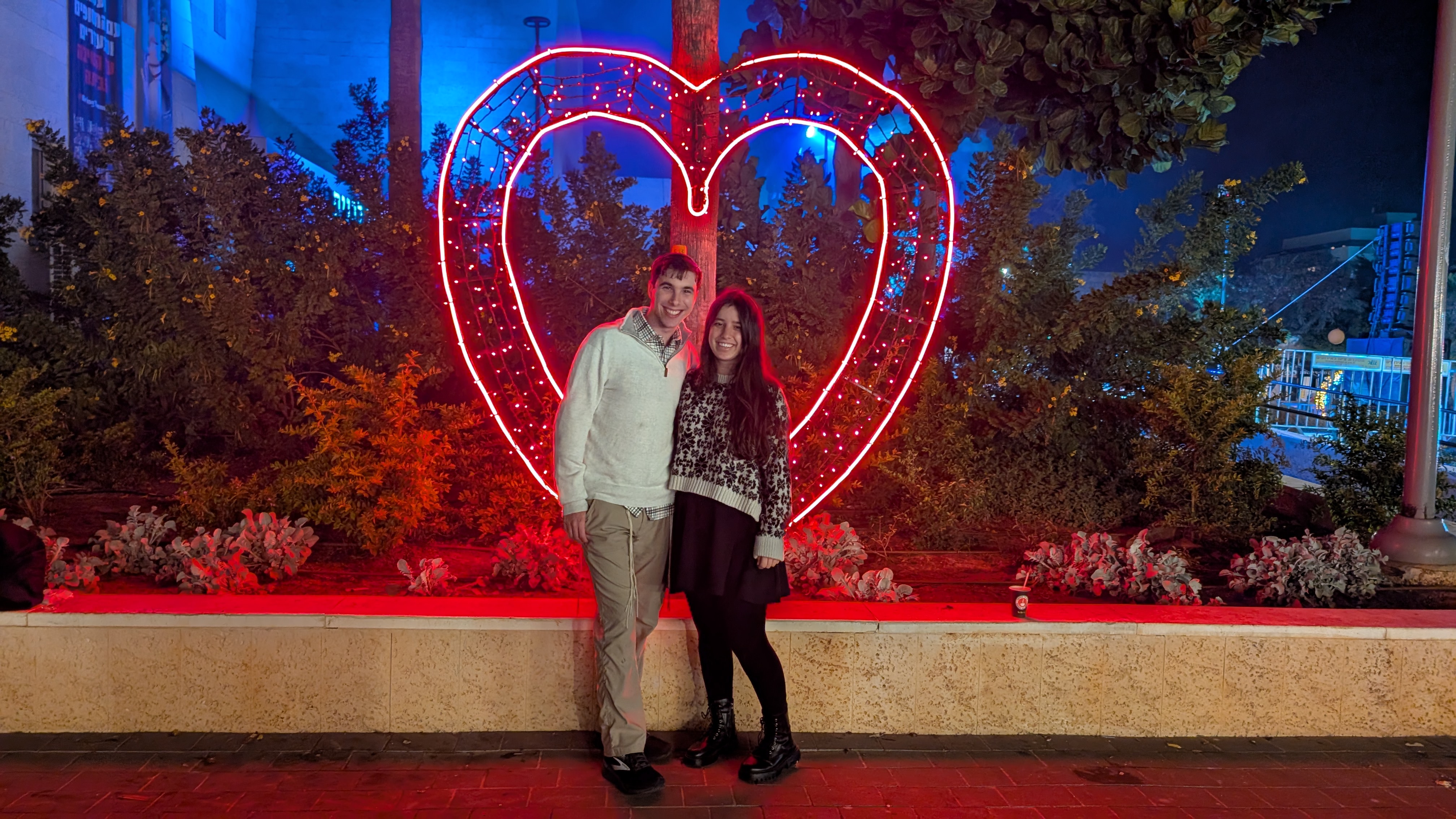 Hershel in cream half-zip sweater and khaki pants, Elia in patterned cream sweater and black skirt, standing in front of a large illuminated red neon heart sculpture at night.