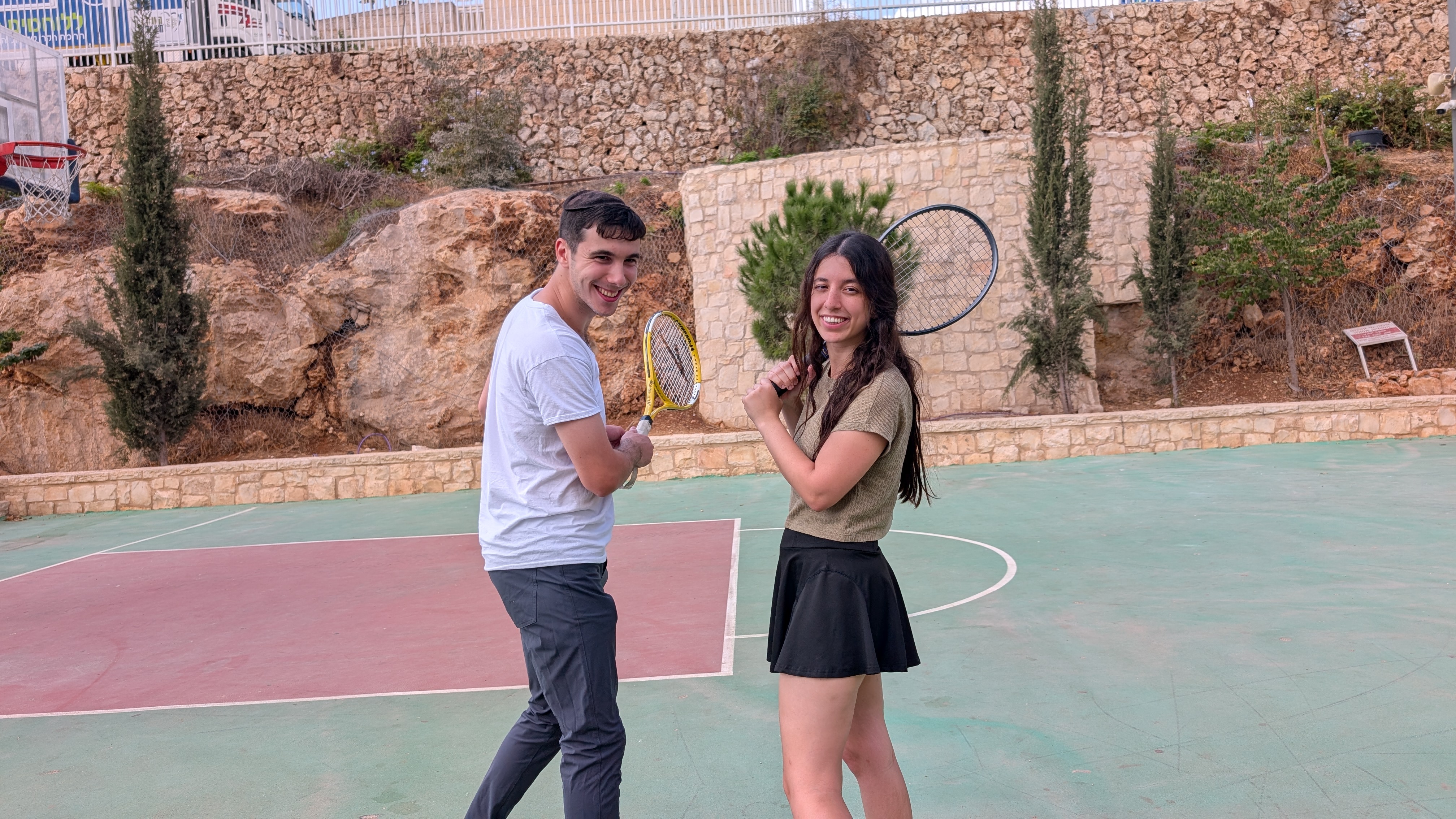 Hershel in white shirt and dark pants, Elia in olive t-shirt and black skirt, on an outdoor sport court with stone retaining wall and trees behind, both in a playful pose with rackets cocked over their shoulders, looking back at the camera.