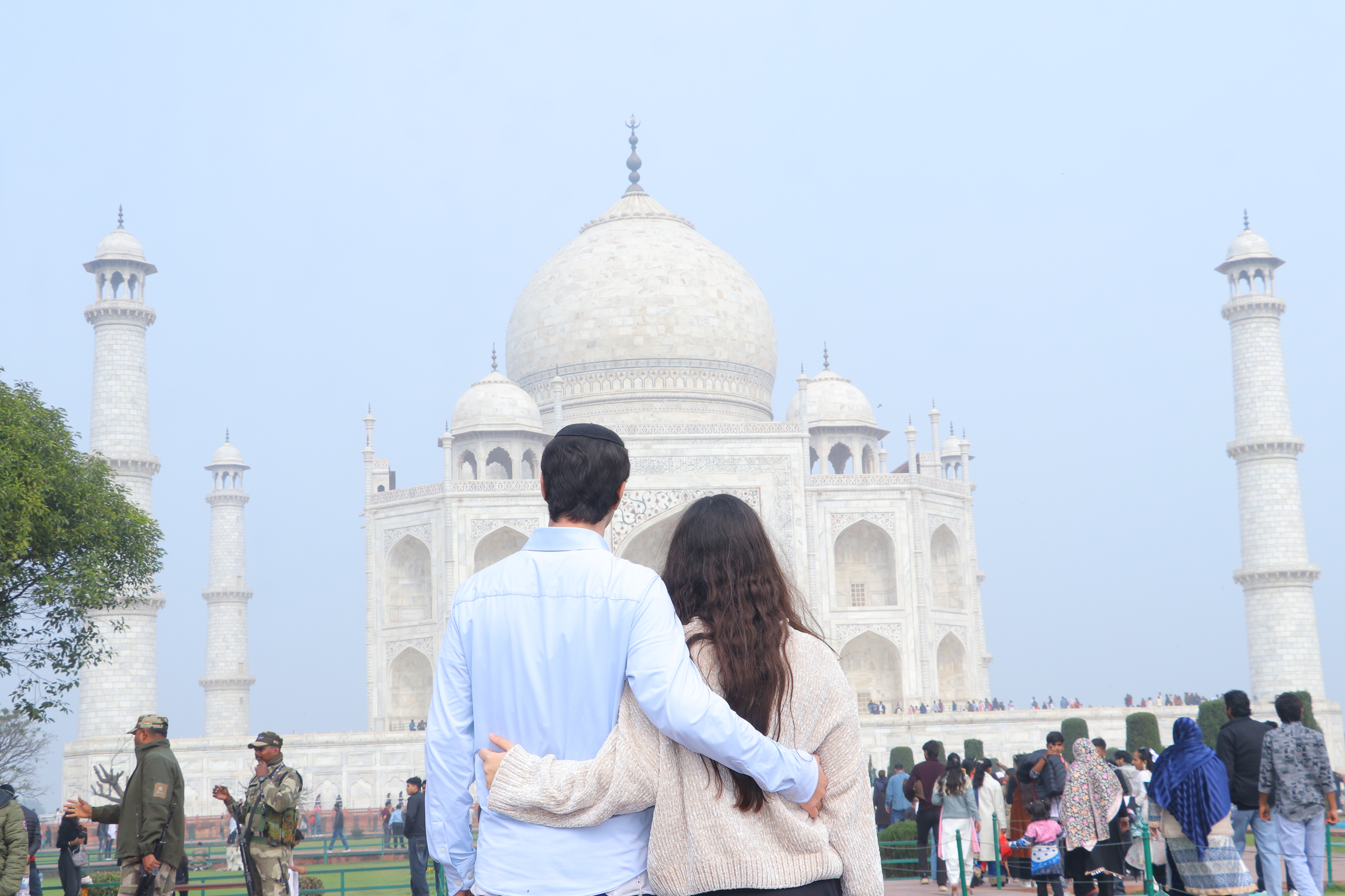 Hershel in light blue dress shirt with kippah and Elia in beige knit sweater with long dark hair, photographed from behind with arms around each other, looking at the Taj Mahal centered in the composition in morning mist.