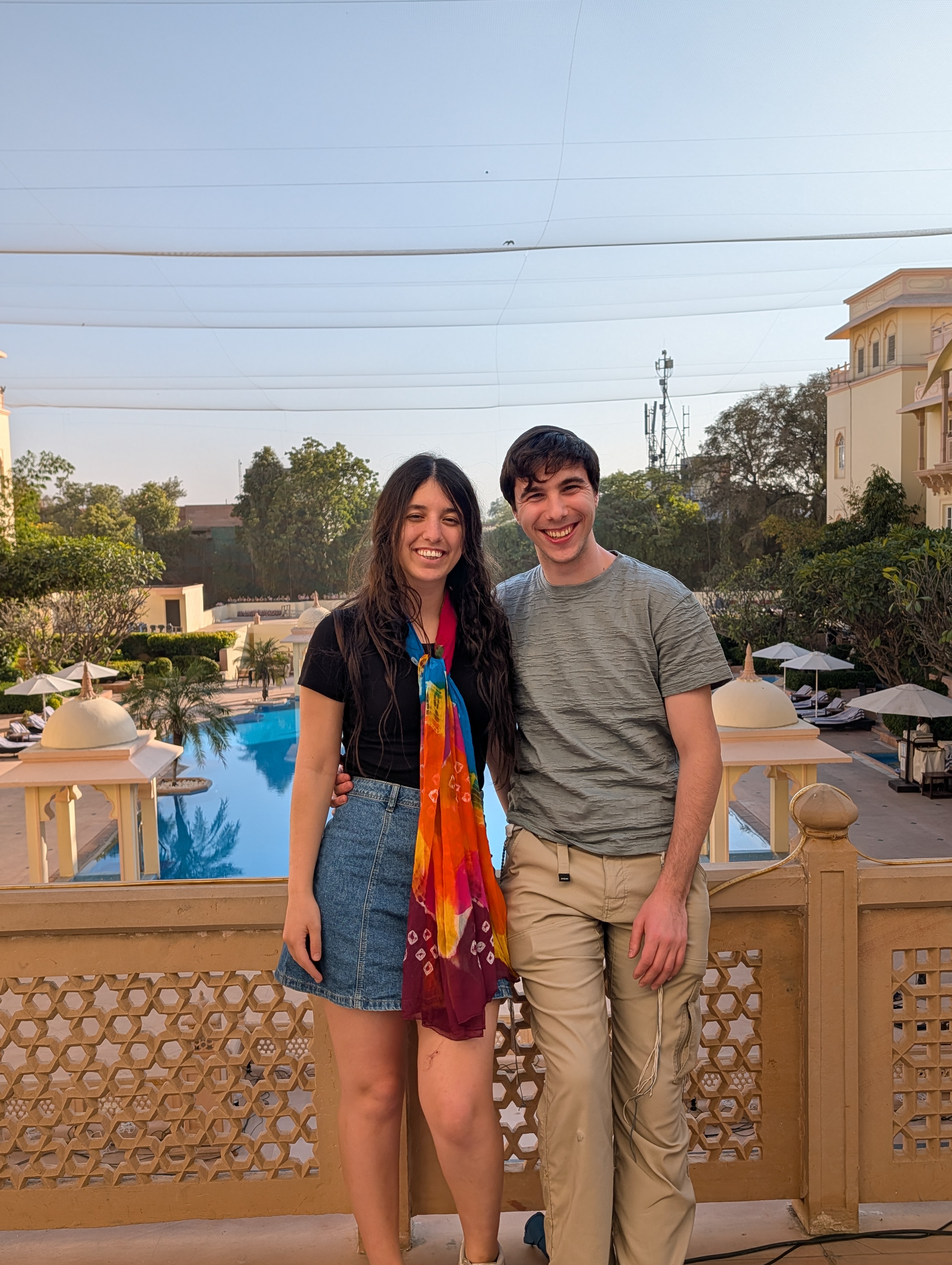 Hershel in olive t-shirt and khaki pants, Elia in black t-shirt and denim skirt with a vibrant tie-dye scarf in red, orange, blue, yellow, and pink, standing together on a hotel balcony in Jaipur with a tiered pool, white pavilions, palm trees, and pink-toned colonial buildings behind them.